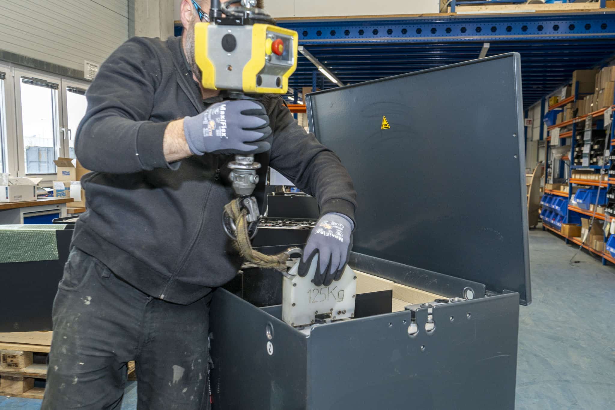 Mitarbeiter bei AKKU SYS hebt mit einem Kranmodul ein 125 kg schweres Batterieelement in ein offenes schwarzes Batteriegehäuse.