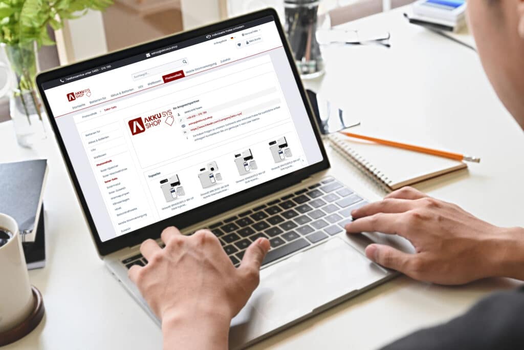 Person nutzt einen Laptop und besucht den AKKU SYS Online-Shop. Auf dem Bildschirm sind verschiedene Produkte für mobile Stromversorgung und Photovoltaik-Komponenten zu sehen.