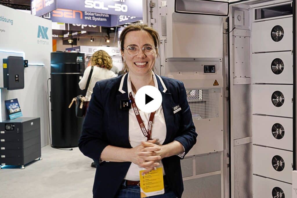Dr. Eva Prumbohm, technische Leiterin von AKKU SYS, steht lächelnd auf einer Messe vor dem geöffneten SAJ Gewerbespeicher CHS2. Im Hintergrund sind weitere Messestände und Besucher zu sehen. Ein weißes Play-Symbol in der Bildmitte deutet auf ein Video hin.
