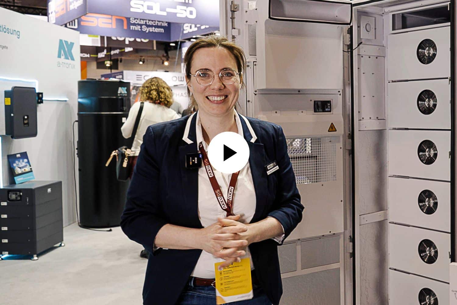 Dr. Eva Prumbohm, technische Leiterin von AKKU SYS, steht lächelnd auf einer Messe vor dem geöffneten SAJ Gewerbespeicher CHS2. Im Hintergrund sind weitere Messestände und Besucher zu sehen. Ein weißes Play-Symbol in der Bildmitte deutet auf ein Video hin.